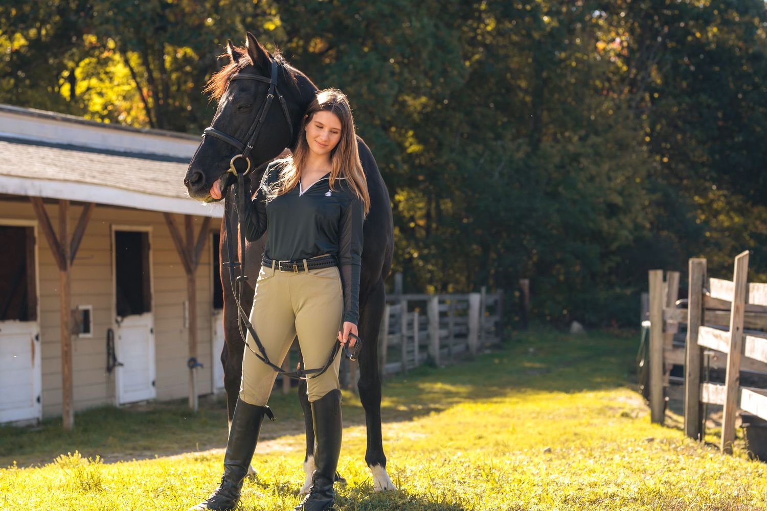 Young woman in equestrian attire standing beside a dark horse near a stable surrounded by trees.