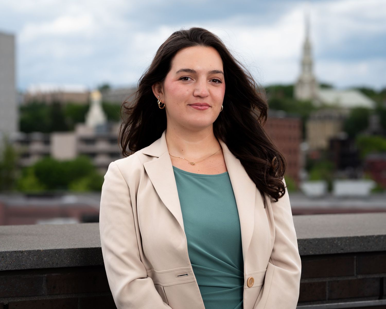 Professional woman in blazer poses on rooftop with city skyline and church steeple in background.