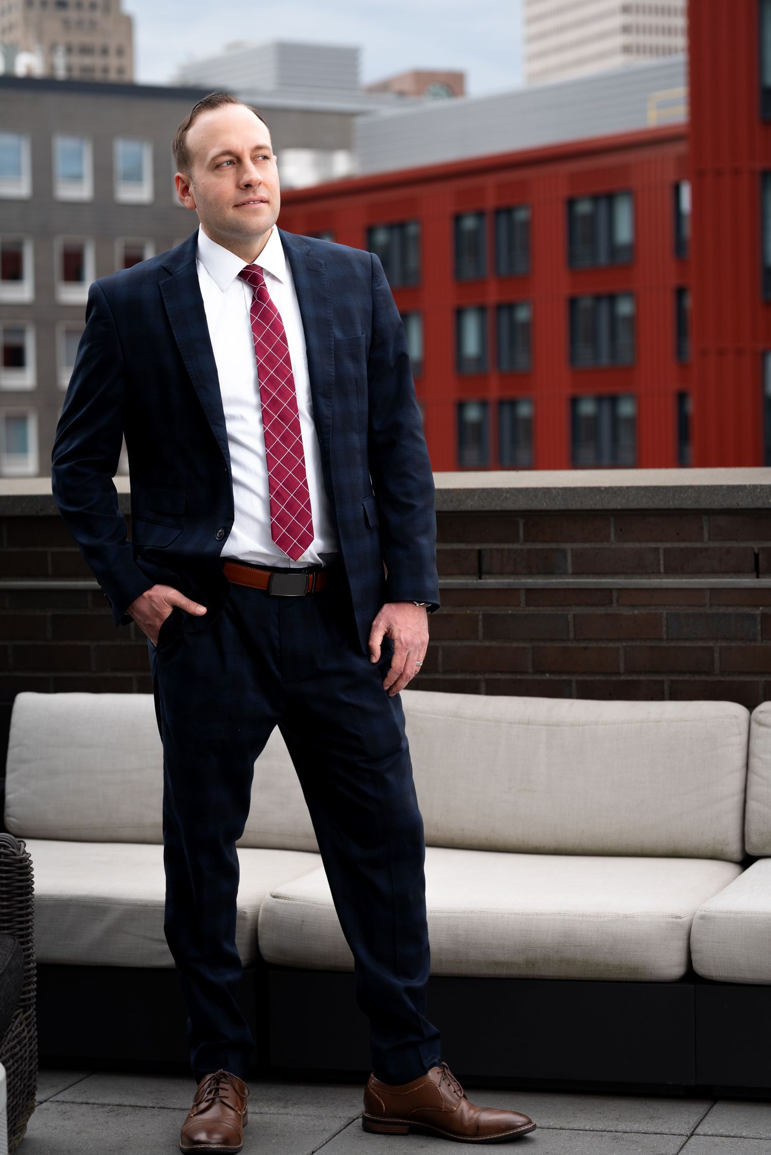 Professional man in navy suit and red tie posing on urban rooftop with city skyline behind him.