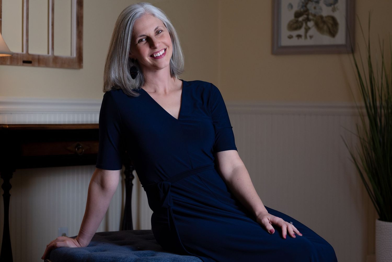 A smiling woman with gray hair wearing a navy blue dress, seated elegantly in a warmly lit room.