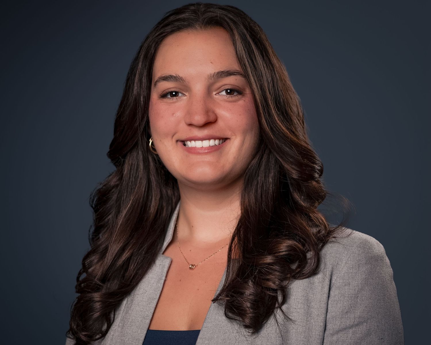 Professional young woman with long brown hair smiling in a gray blazer for a business headshot.