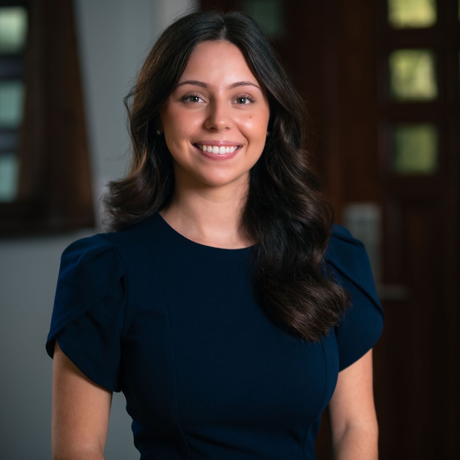 Young professional woman with dark wavy hair smiling in a navy blue dress in a warmly lit interior.