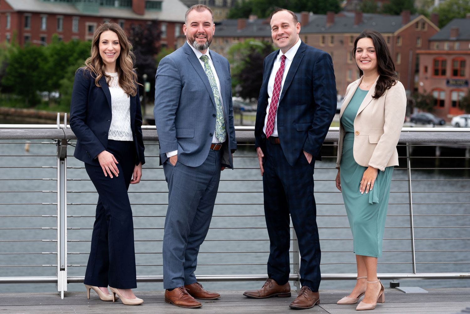 Four business professionals smiling outdoors near a waterfront, dressed in formal attire on a sunny day.