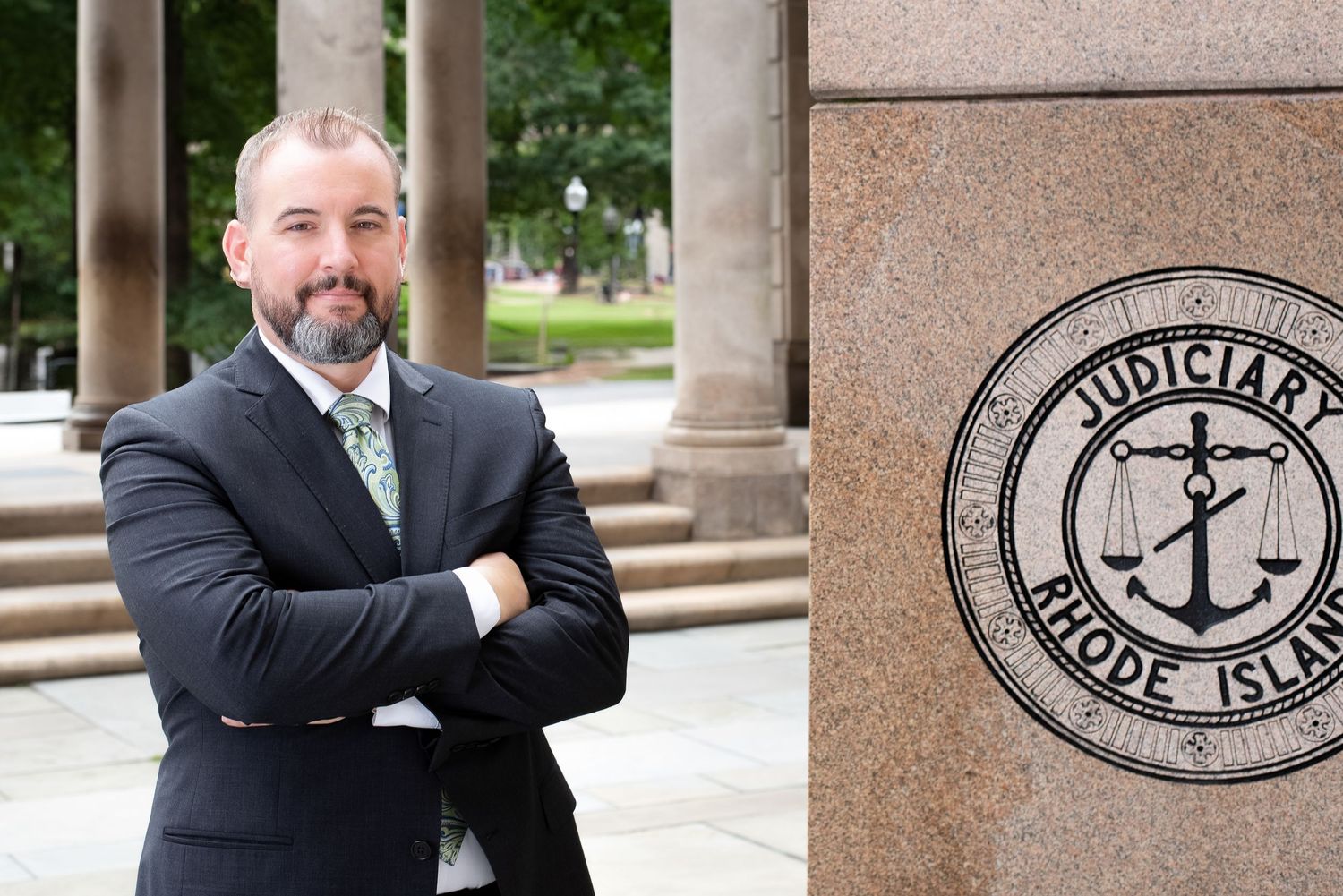 Man in suit stands arms crossed near Rhode Island Judiciary seal on stone pillar outside courthouse.