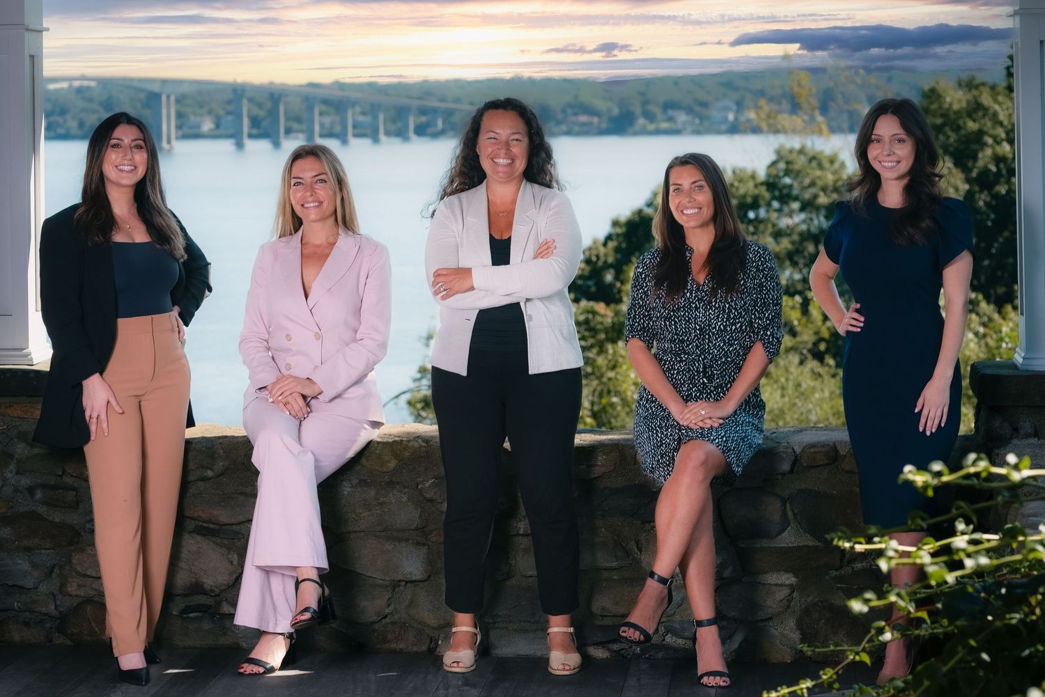 Five professional women posing together outdoors with a scenic river and bridge backdrop at sunset.