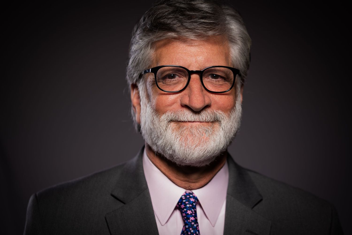 A distinguished older man with gray beard and glasses smiles warmly wearing a suit and tie.