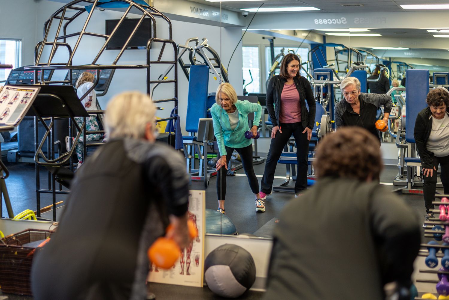 Group fitness class with women exercising in a modern gym using medicine balls and various workout equipment.
