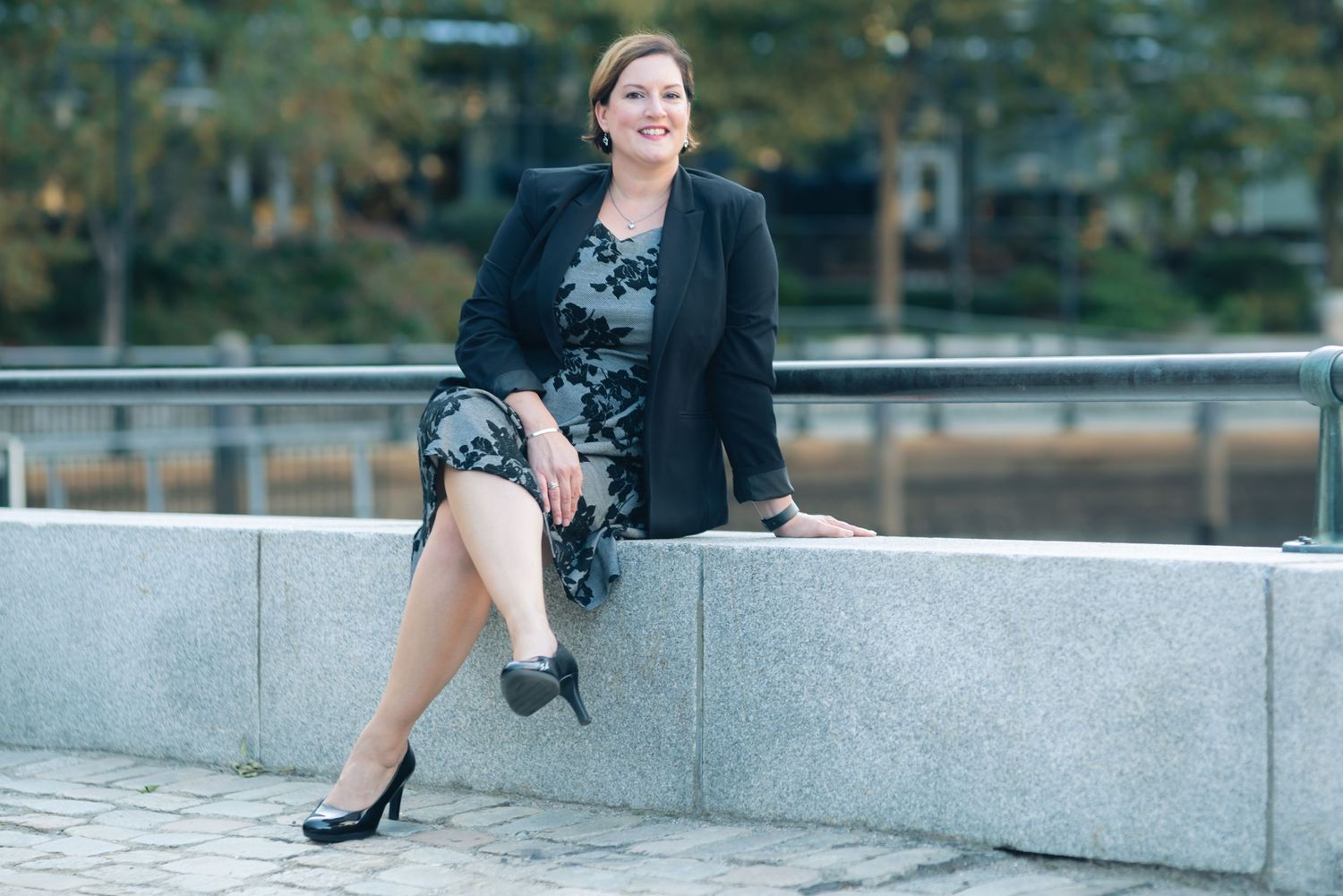 Professional woman in black blazer and floral dress sitting on stone wall in urban setting.