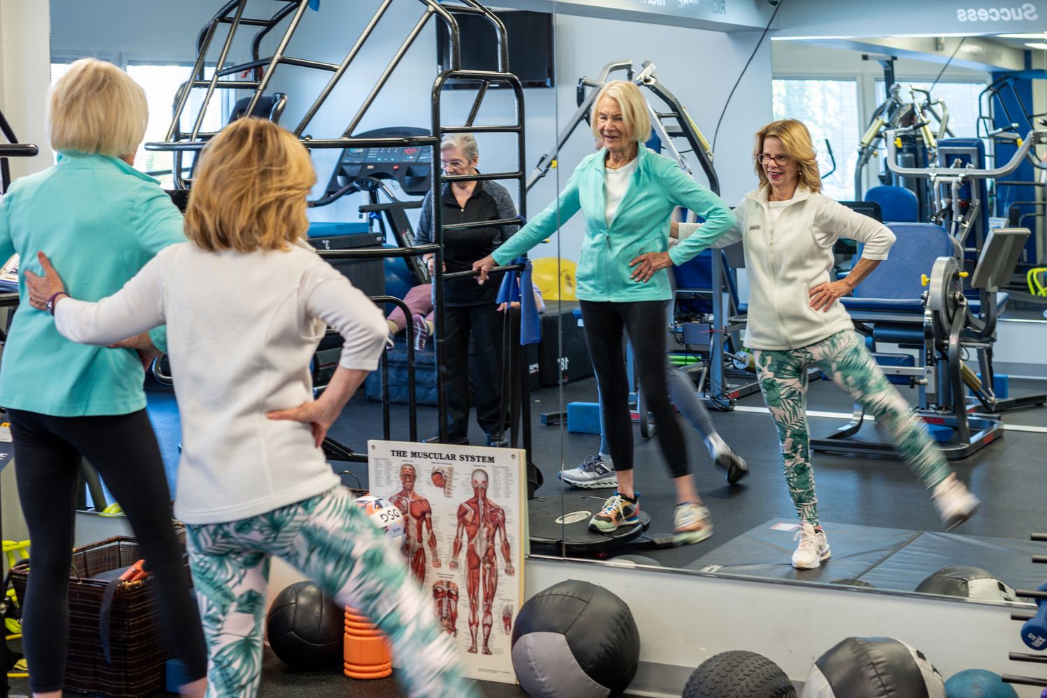 Women exercising in a gym, doing aerobics near mirrors and fitness equipment, wearing teal and white workout clothes.