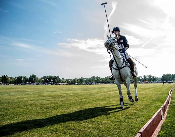 USA polo player riding a white horse along the field boundary in bright sunlight.