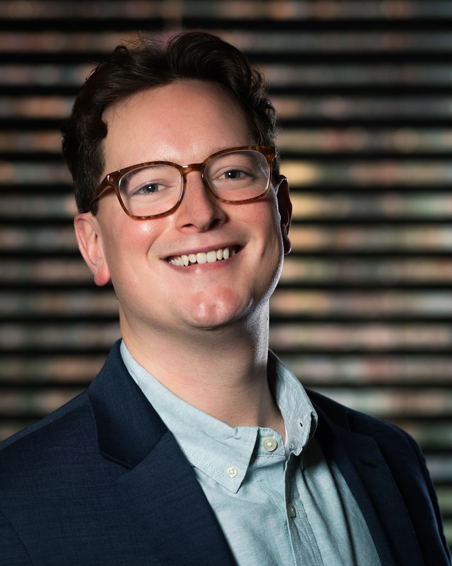 Young smiling man wearing tortoise-shell glasses and a navy blazer with a colorful bokeh background.