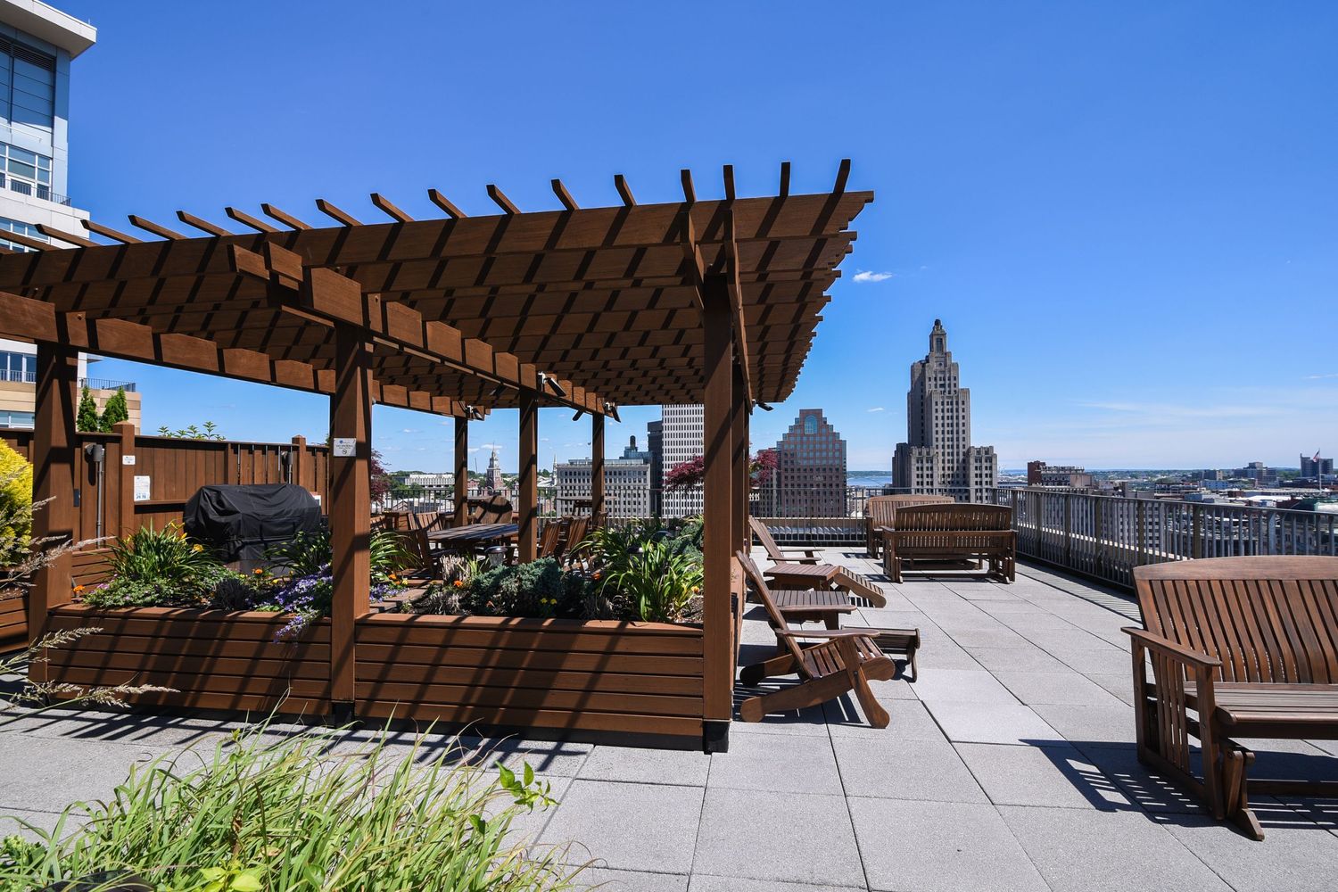 Rooftop terrace with wooden pergola, garden planters, and city skyline views on sunny day.