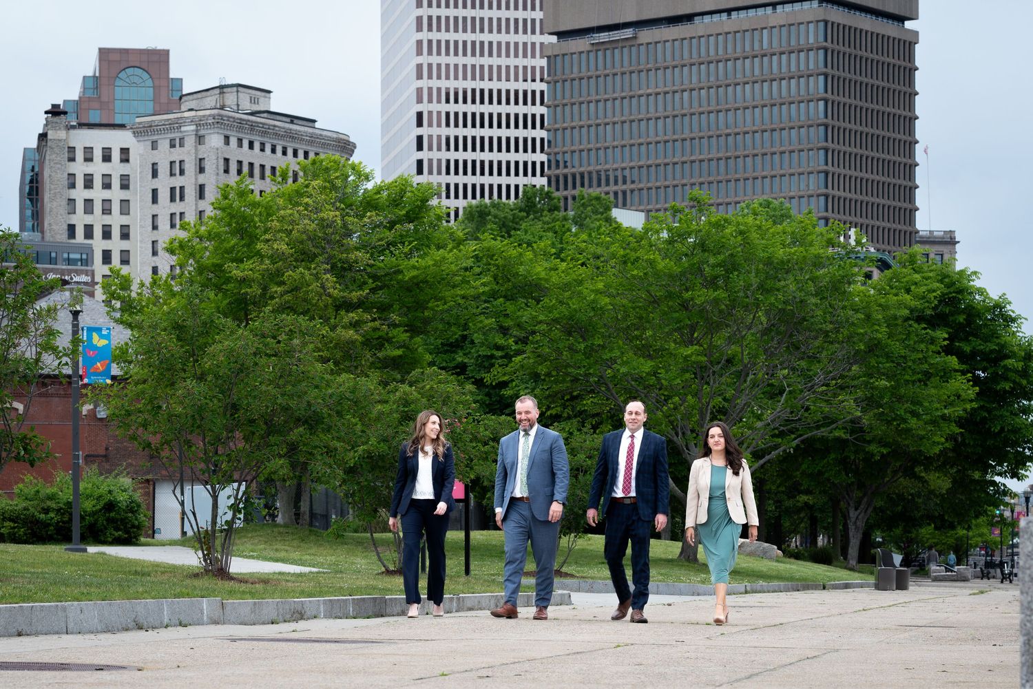 Four business professionals walking outdoors near green trees with city skyscrapers in the background.