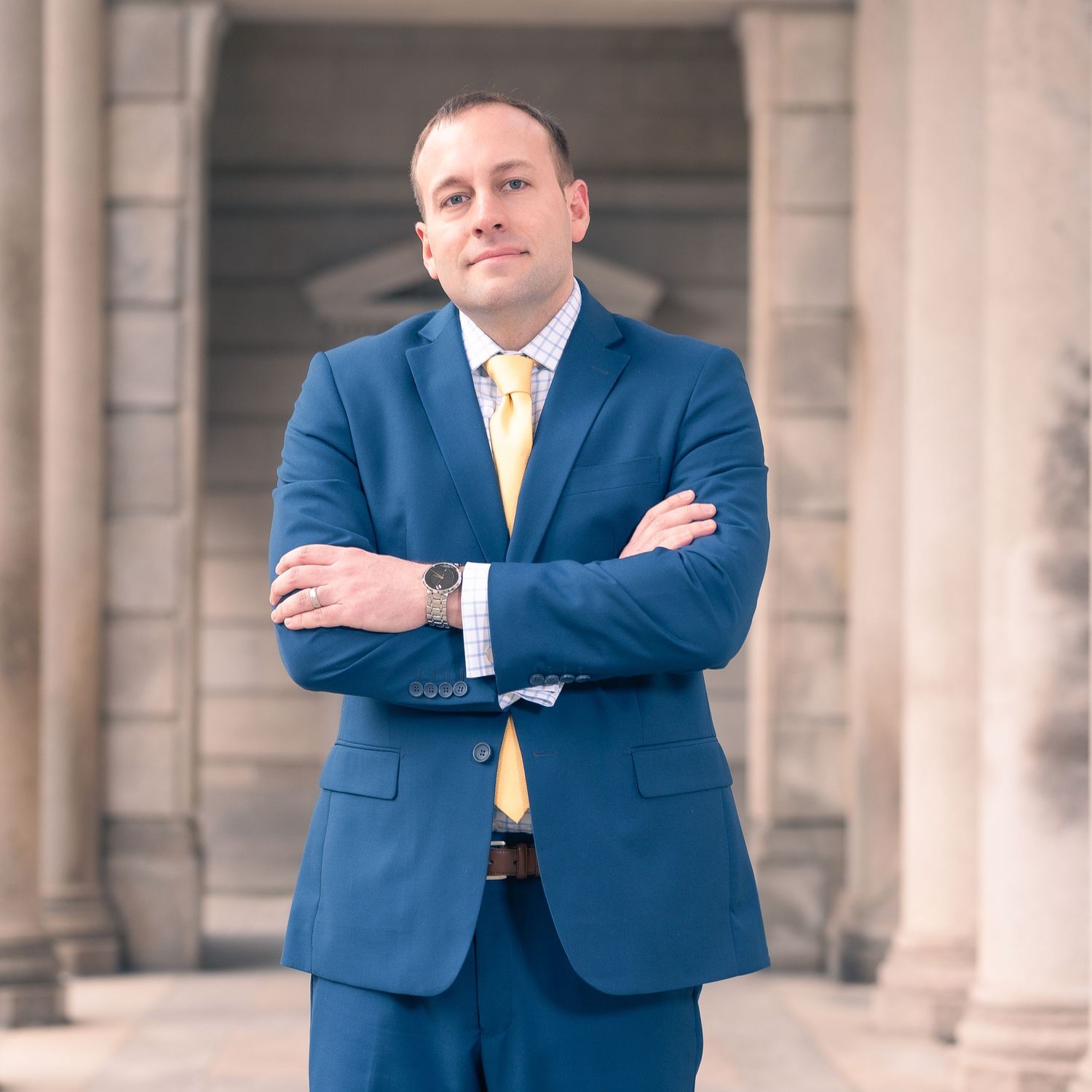 A confident man in a blue suit and yellow tie stands with arms crossed before classical columns.