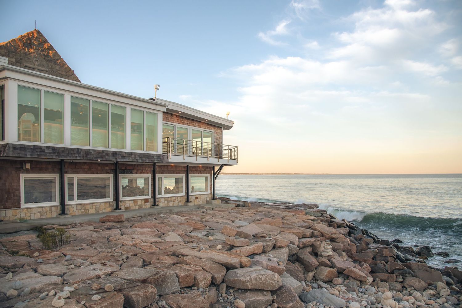 Oceanfront building with large windows sits on rocky shoreline at sunset with calm sea views.