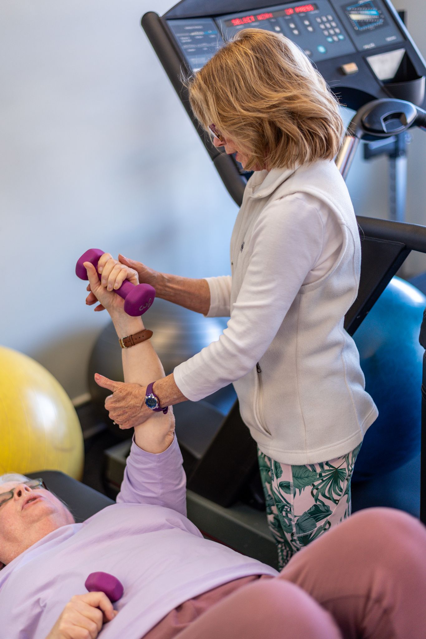 Physical therapist assists senior patient with dumbbell exercise in rehabilitation gym with treadmill and exercise balls.