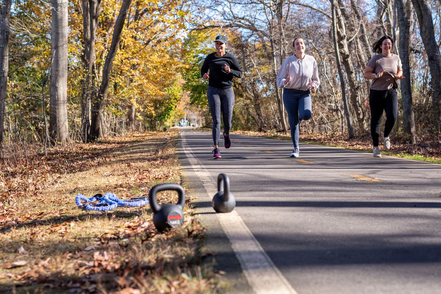 Three runners jogging on a tree-lined road in autumn with kettlebells and a towel on the pavement foreground.