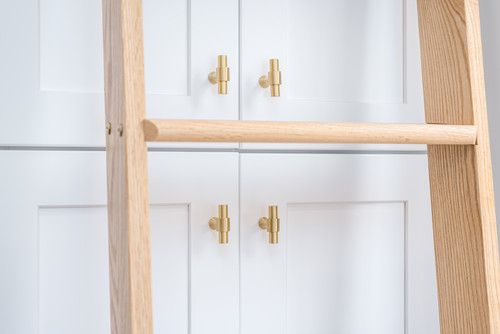 Wooden ladder shelf leaning against white shaker cabinets with gold brass T-bar handles in a modern home.