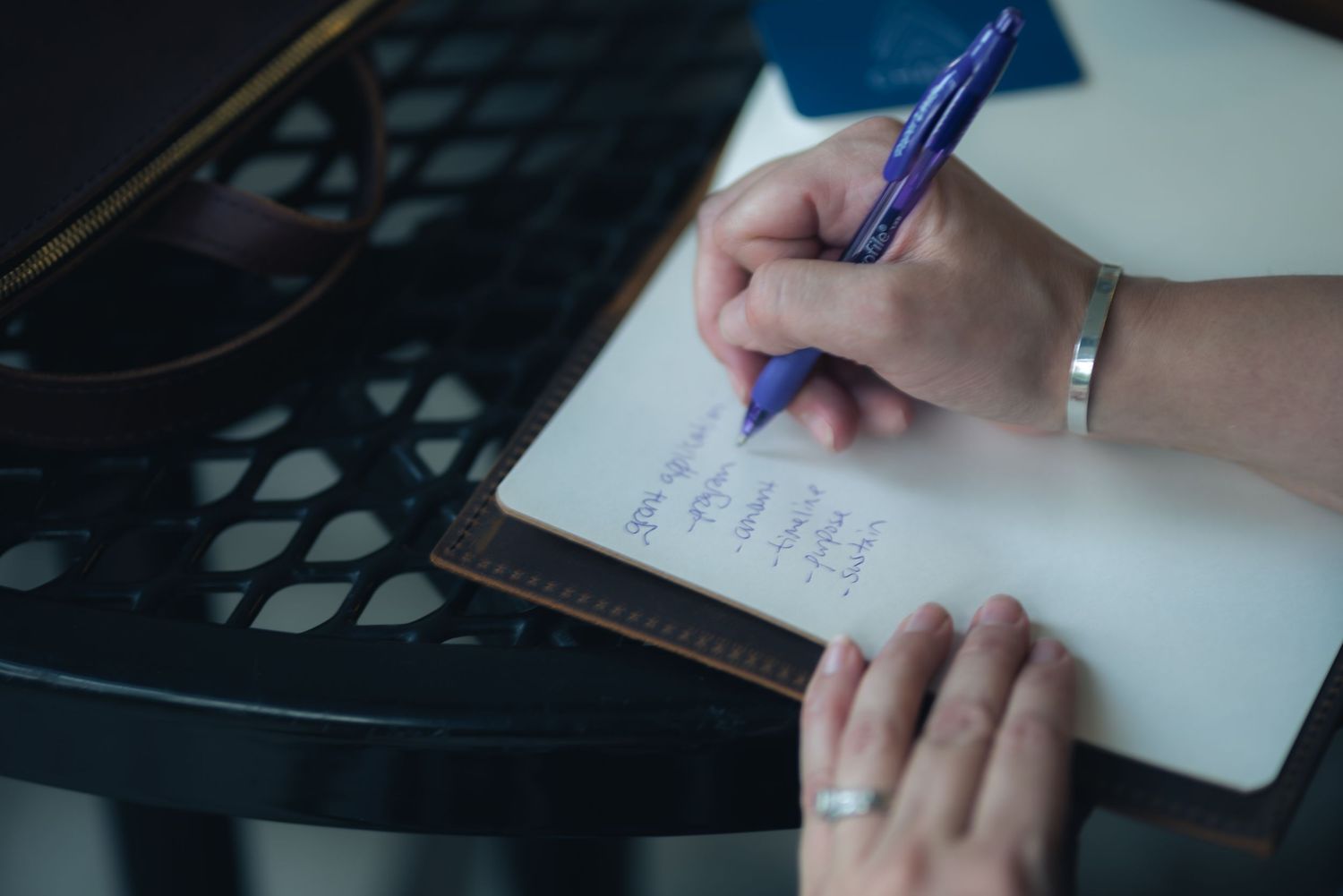 Close-up of a hand writing notes in a notebook with a purple pen on a black table.