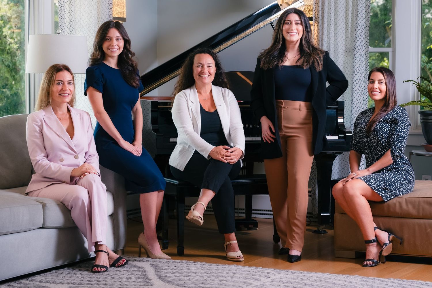Five professional women posing together in an elegant living room with a grand piano.