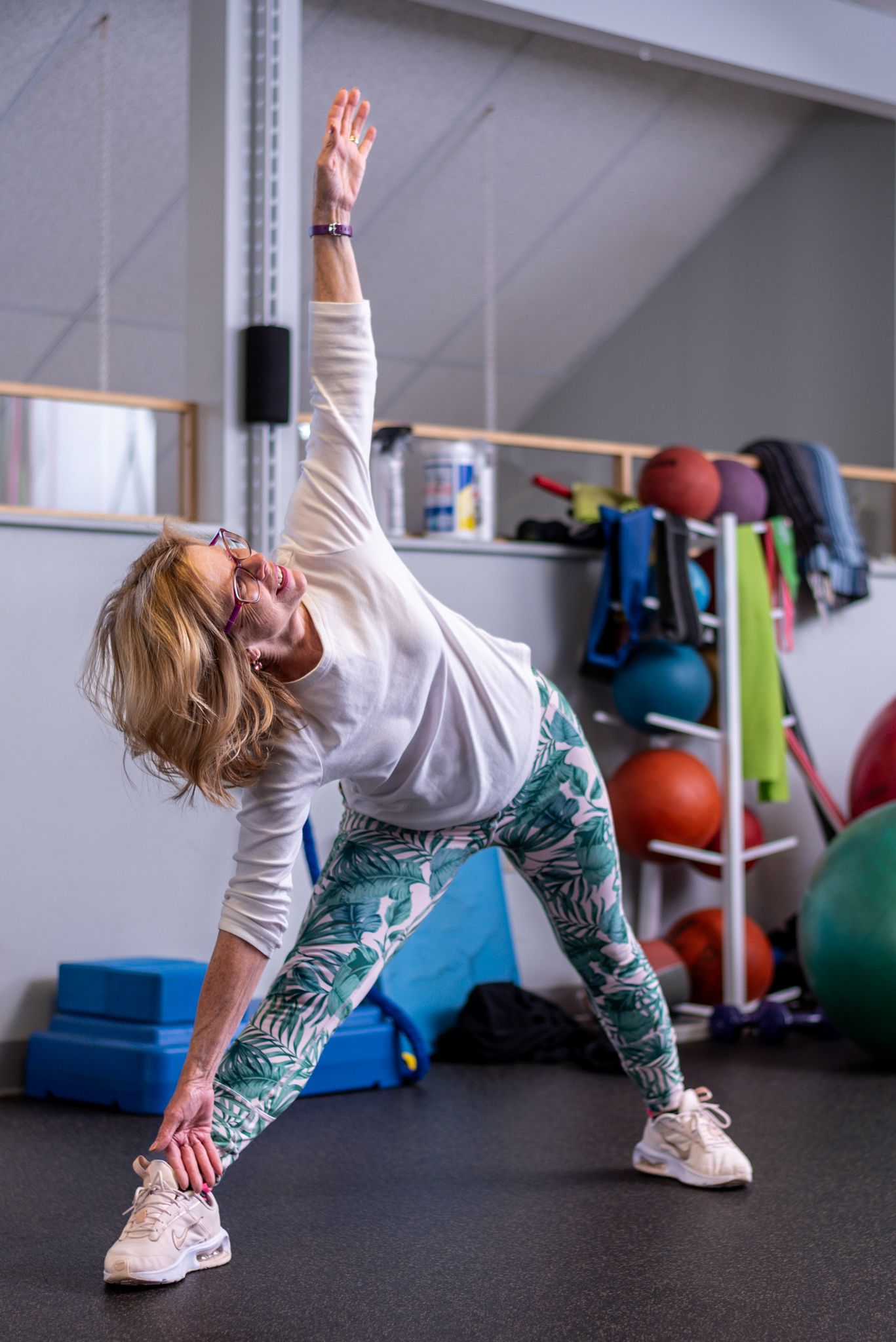 Woman in patterned leggings doing a triangle stretch pose in a gym with colorful exercise balls in background.