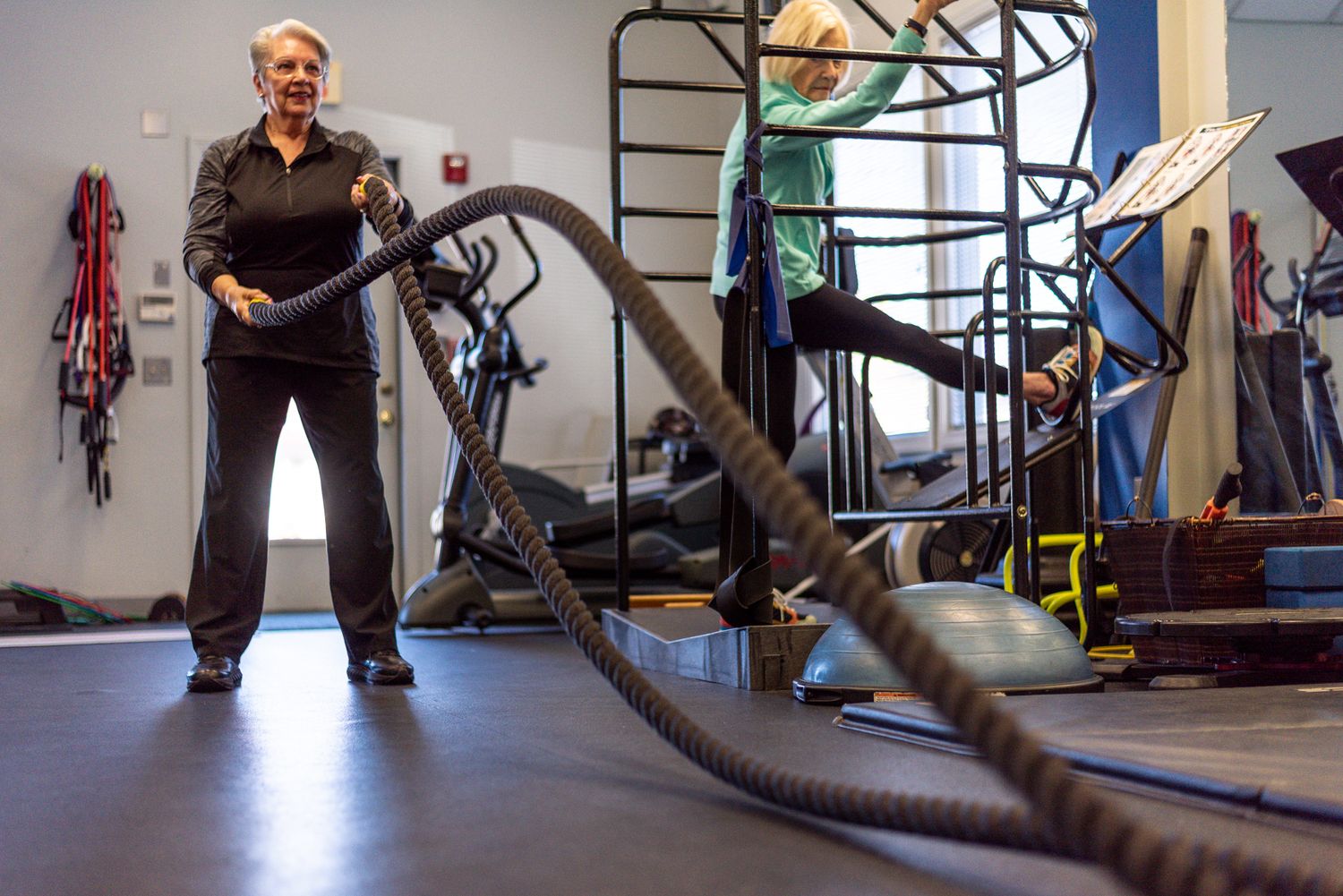 Woman doing battle rope workout exercise in a modern gym with fitness equipment in the background.