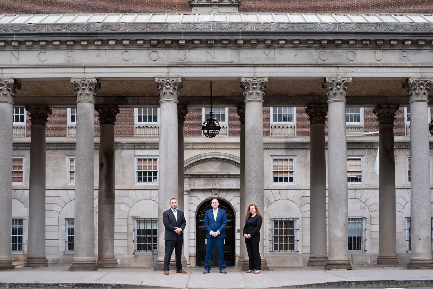 Three professionals in formal attire stand spaced apart before a grand columned brick building.