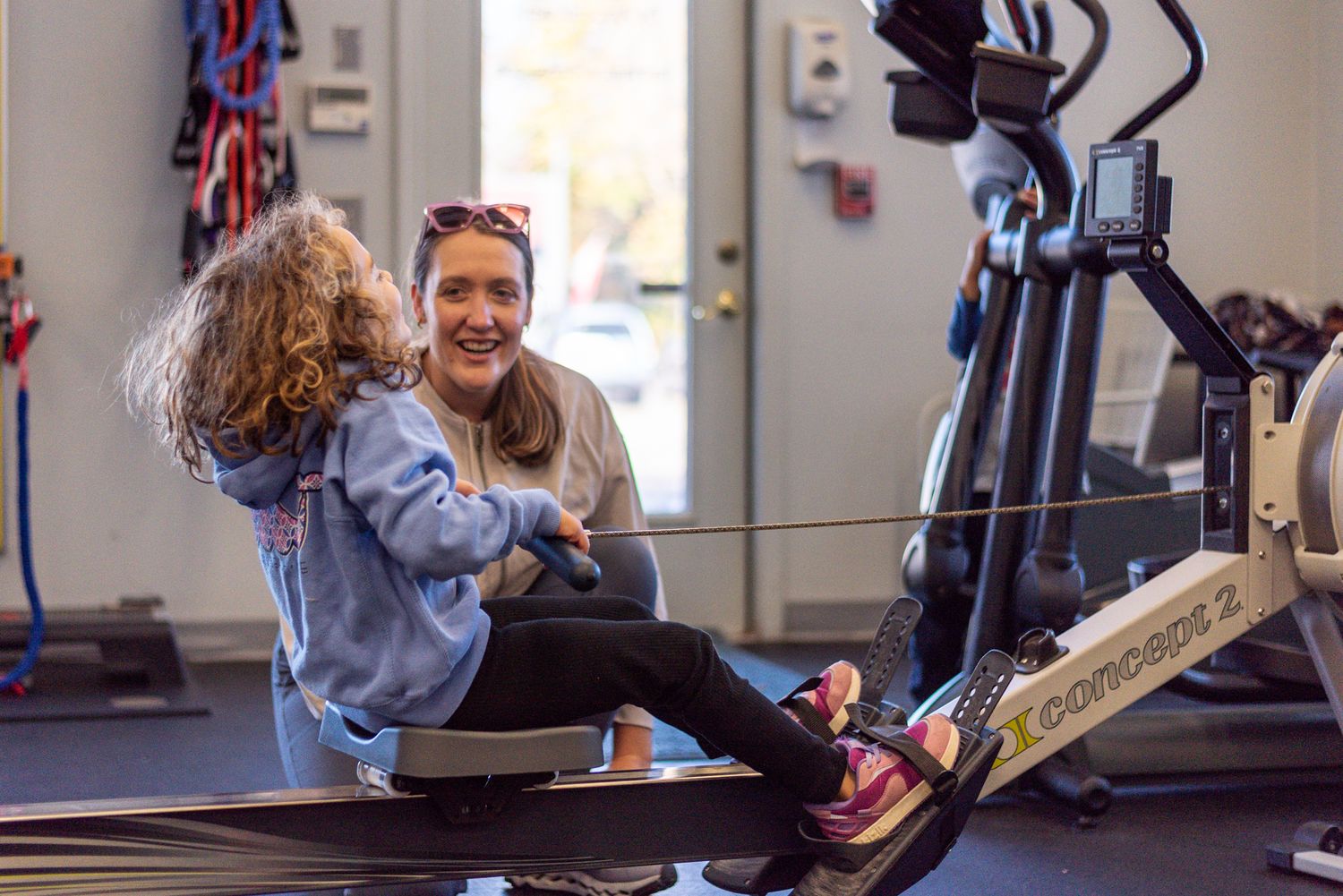 Woman smiling with young girl on Concept2 rowing machine in bright gym setting.