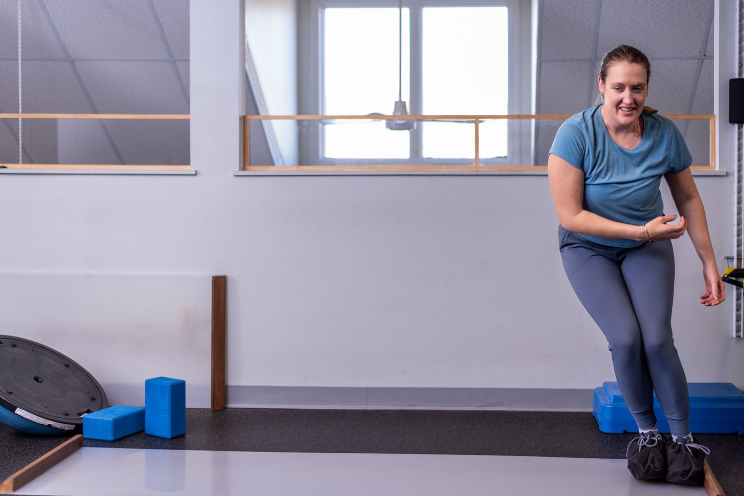 Woman in blue top and gray leggings exercising in a bright gym with blue yoga blocks on the floor.