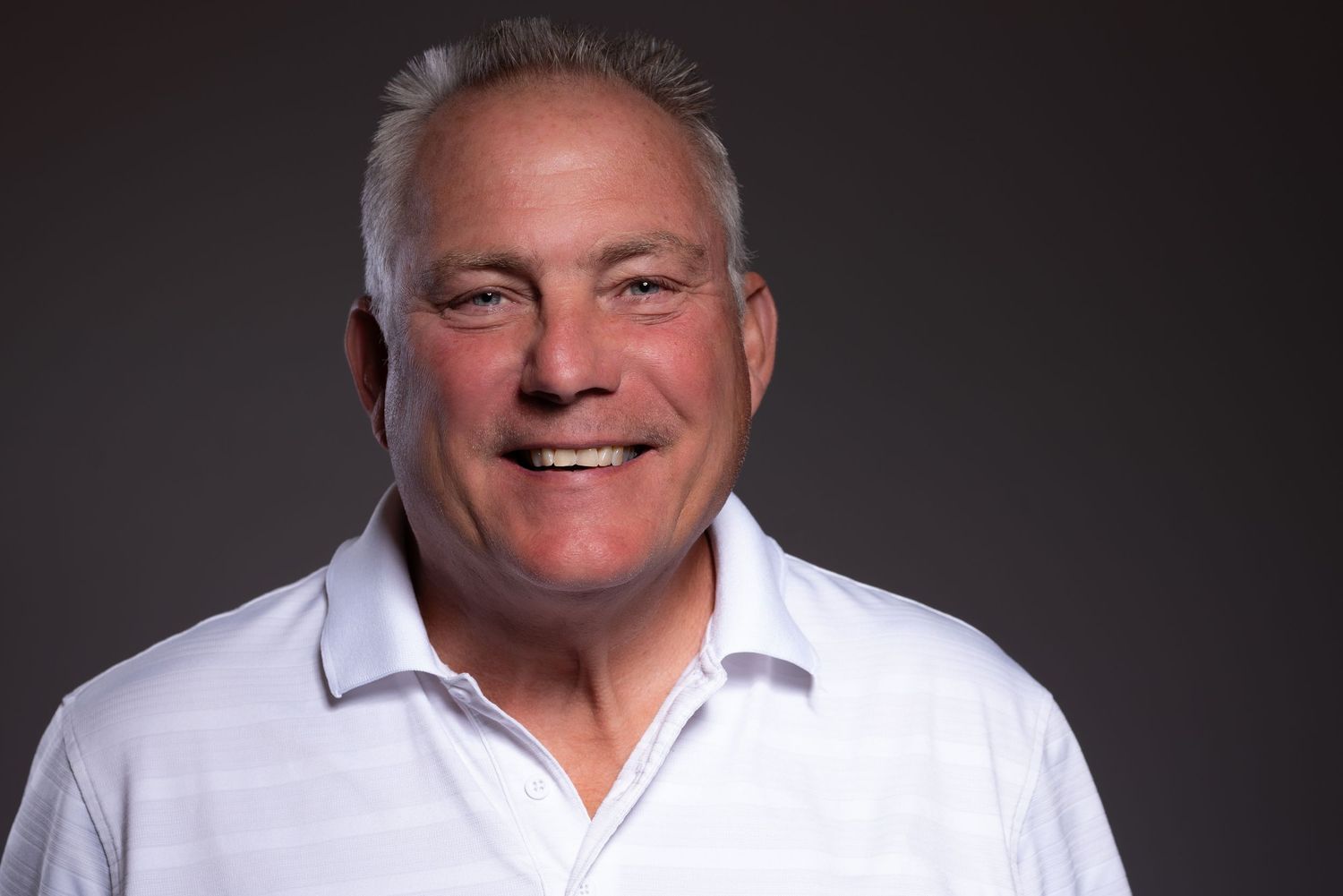 Smiling middle-aged man with gray hair wearing a white polo shirt against a dark gray studio background.