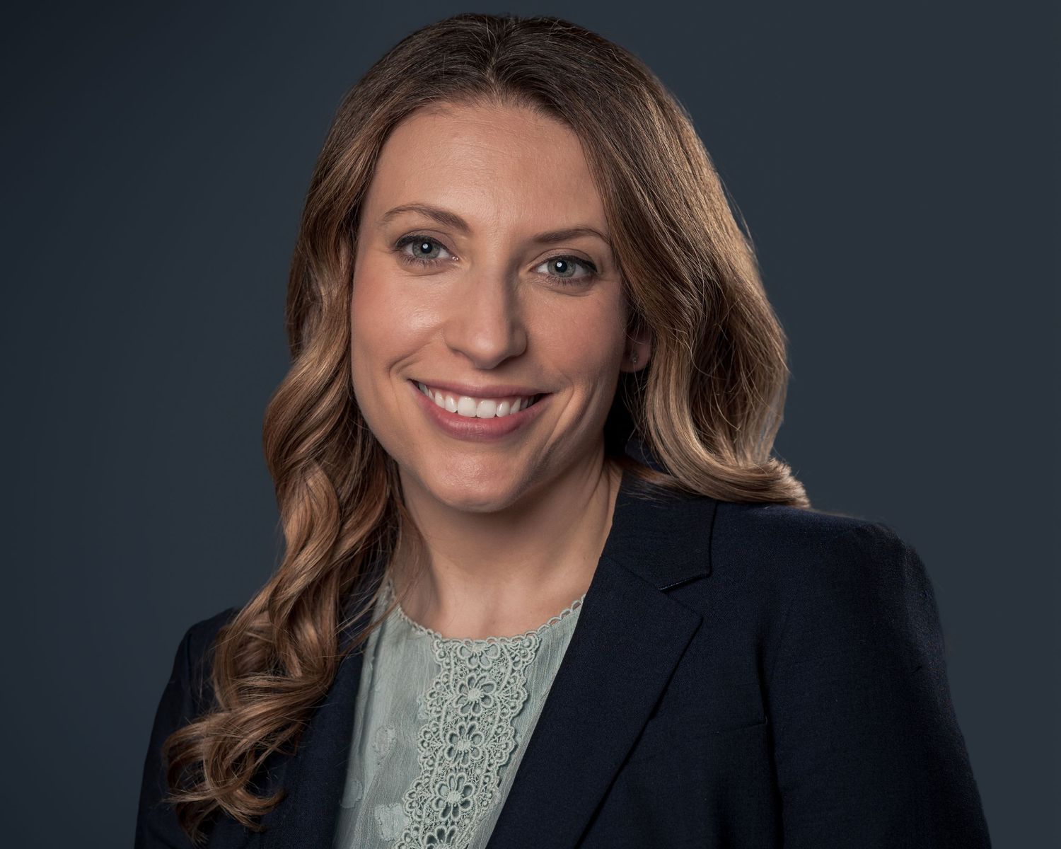Professional headshot of a smiling woman with wavy hair wearing a navy blazer and lace blouse against a dark background.