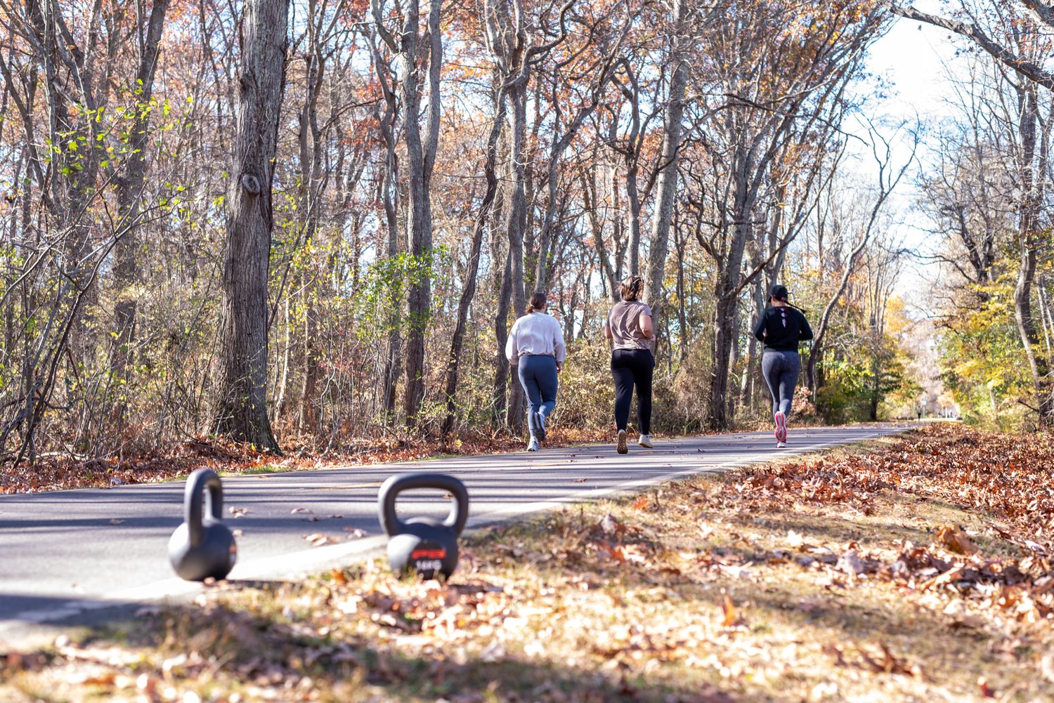 Group of people jogging on a tree-lined path in autumn with kettlebells in the foreground.