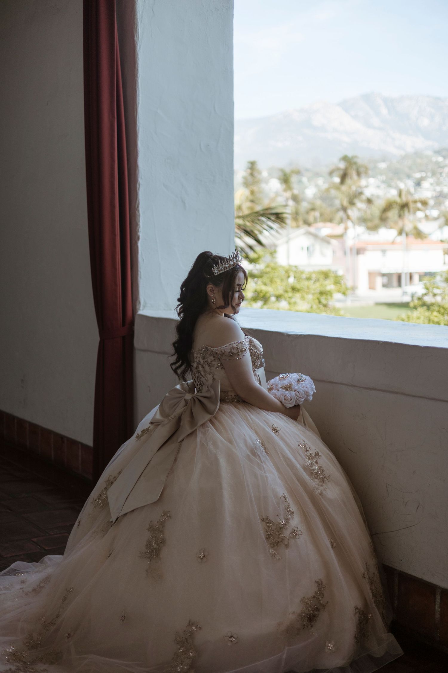Elegant bride in an ornate off-shoulder ball gown looks out window at mountain landscape on her wedding day.