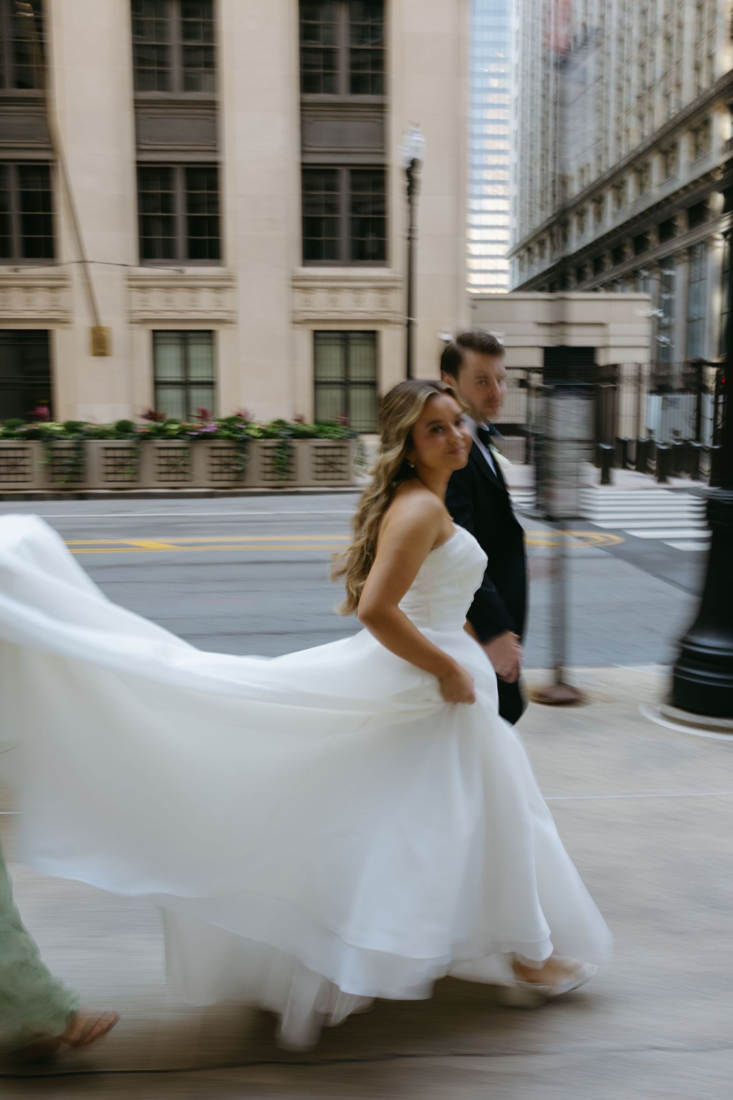 A flowing white wedding dress twirls in motion on a city street as a couple walks past elegant architecture.
