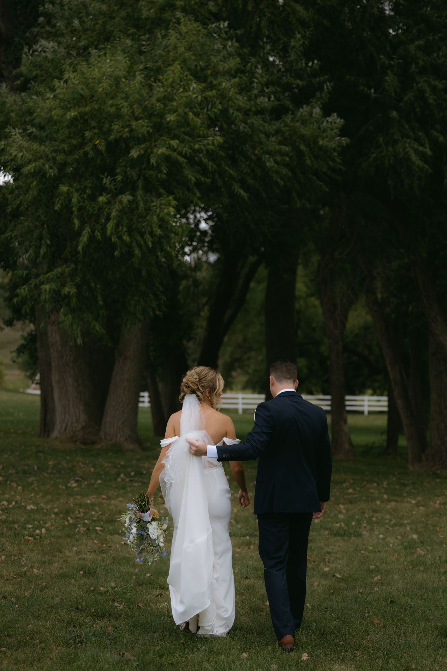 A couple in wedding attire walks through a serene park setting with tall trees and green grass.