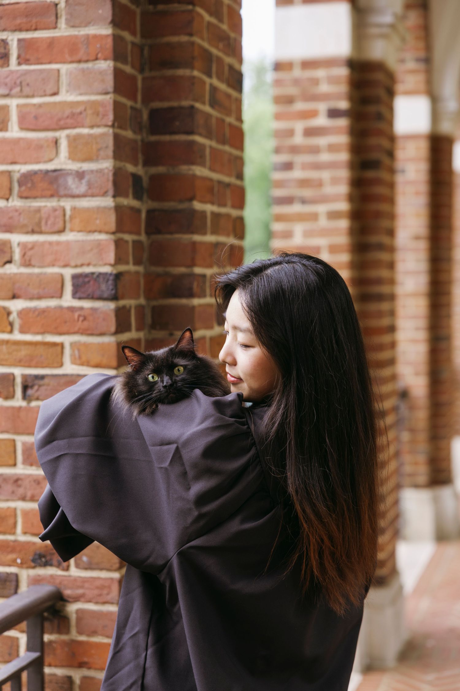 A long-haired woman in a black sweater holds a black cat while standing next to a brick wall.