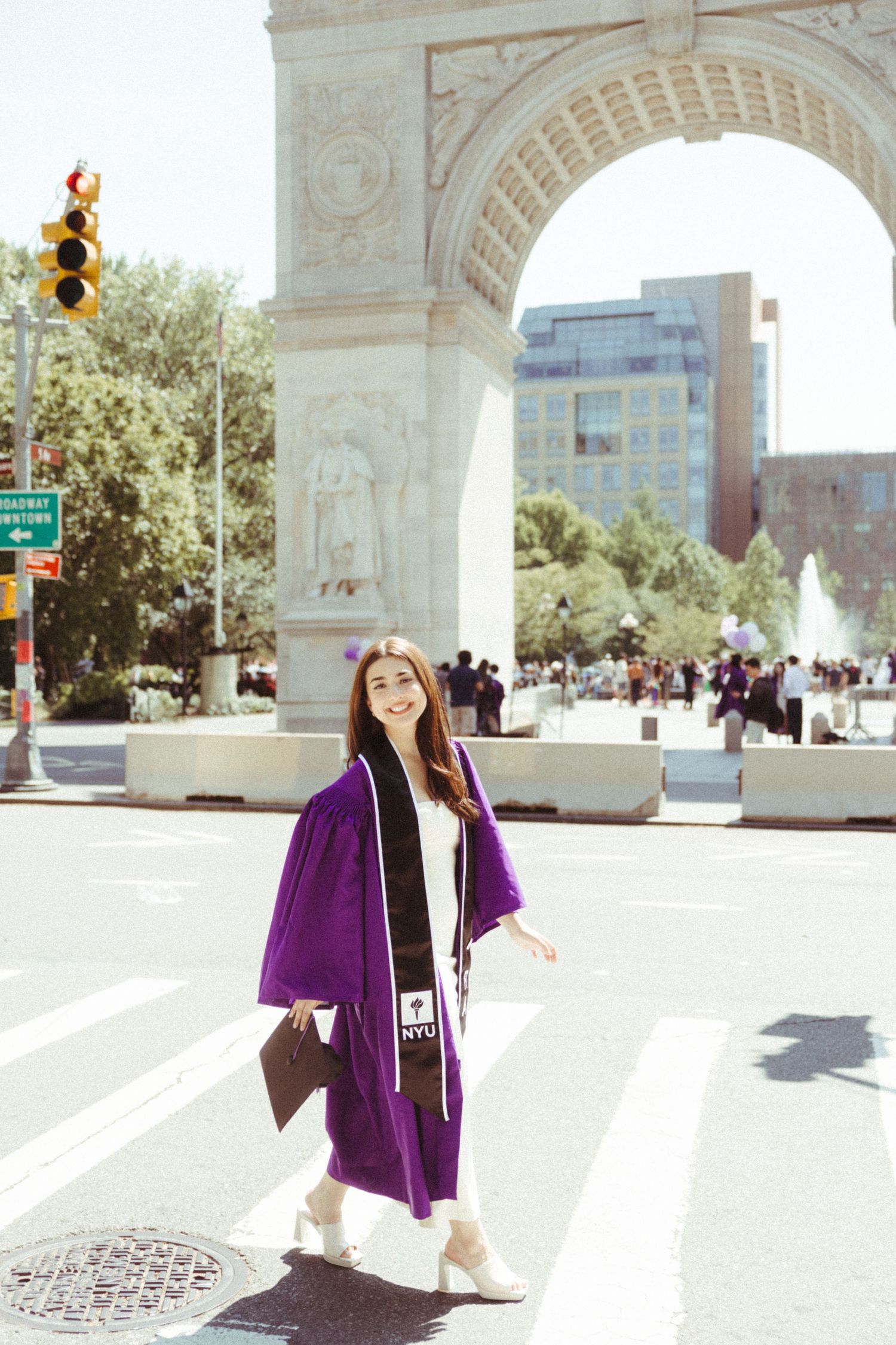 A graduate in a purple graduation gown poses in front of Washington Square Arch on a sunny day.