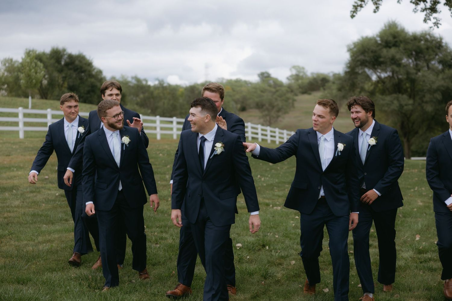 A group in dark suits stands together outdoors on a grassy lawn with white fencing visible in the background.