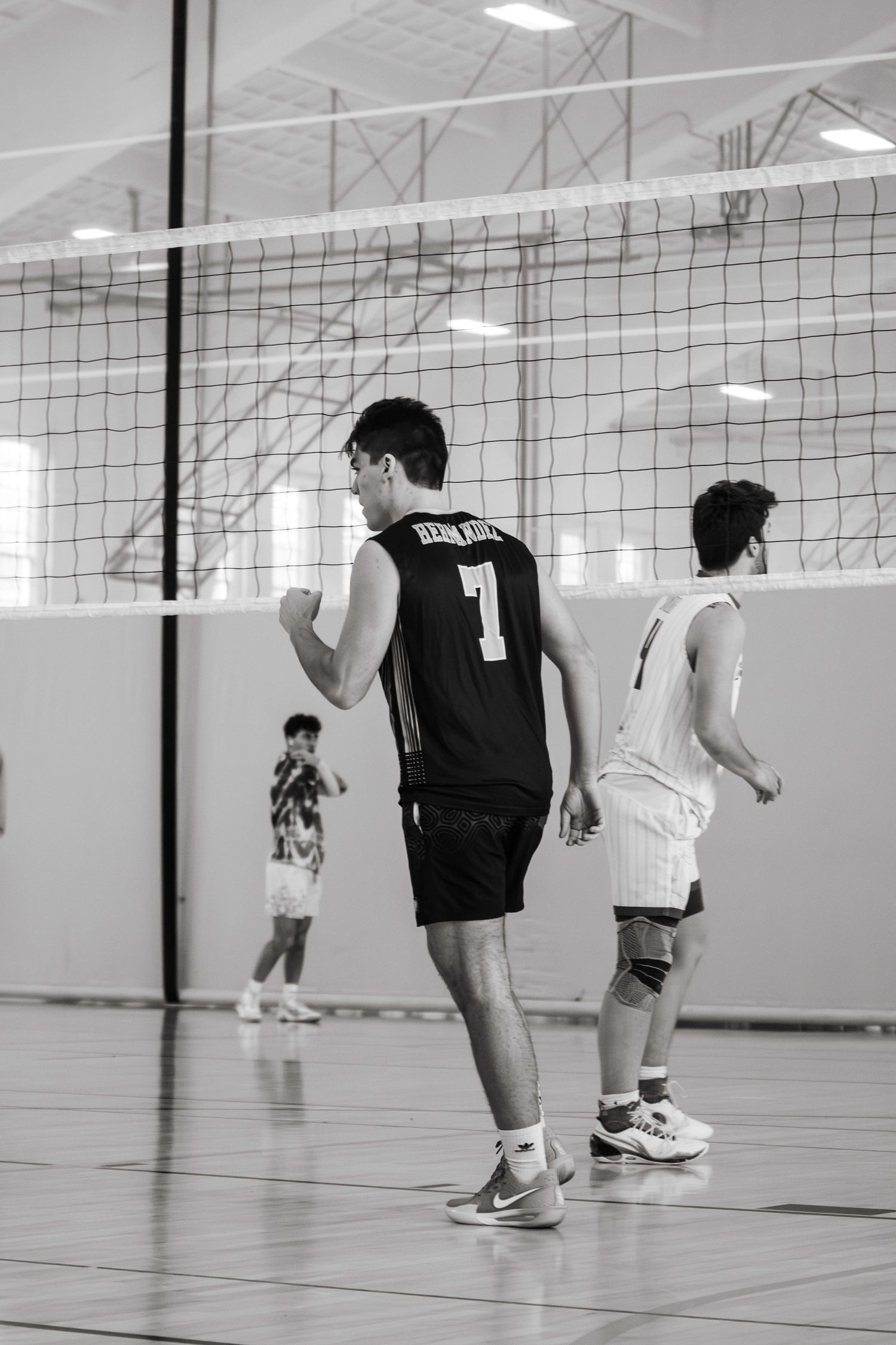 Volleyball players in black and white jerseys stand at the net during an indoor game while a figure watches in background.