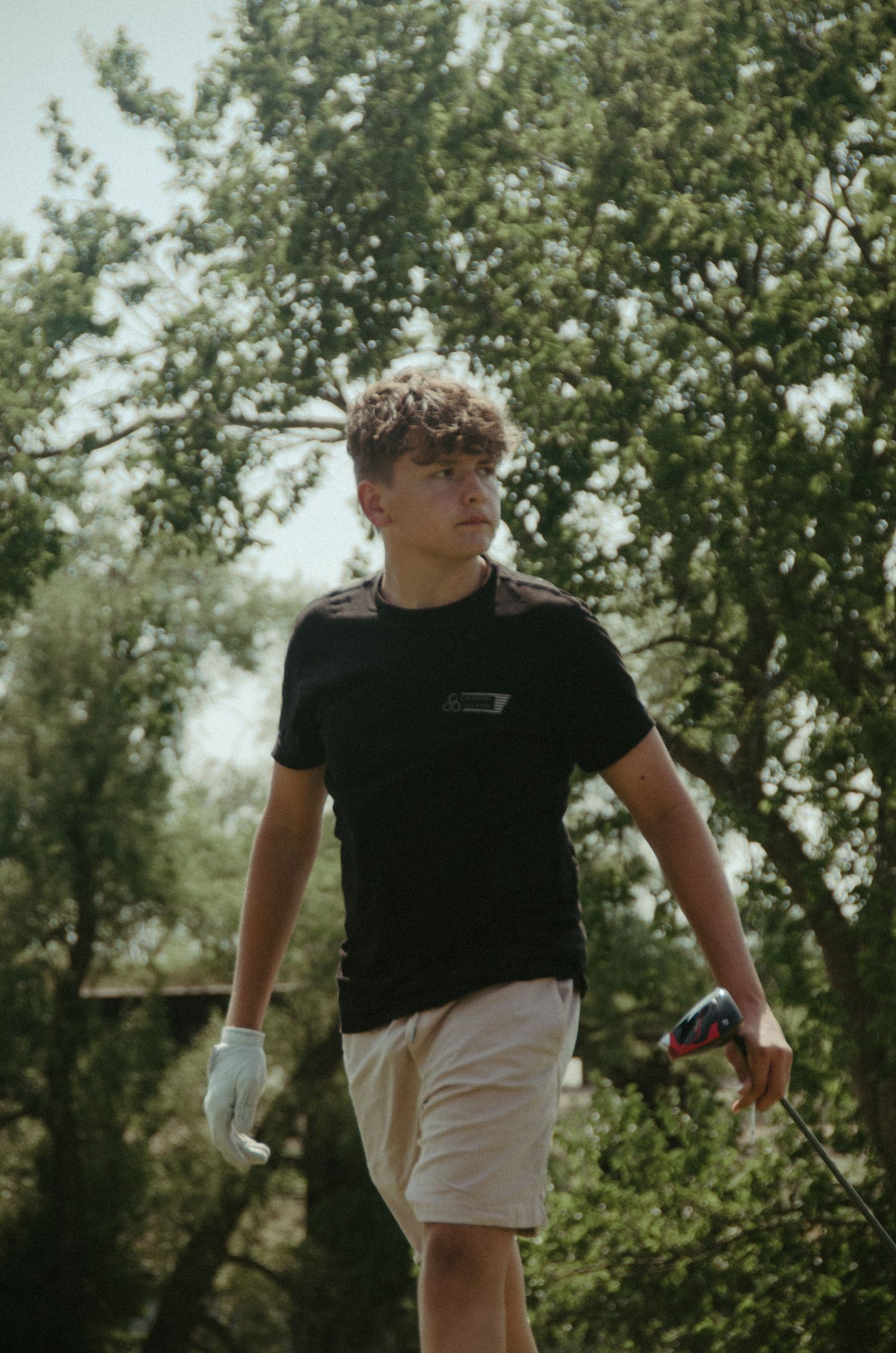 Young golfer in black shirt and white shorts standing among trees on sunny day.