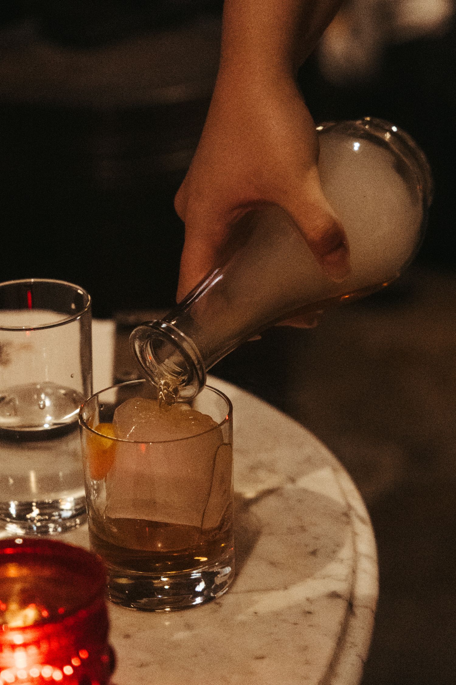 A hand pours amber liquid into a cocktail glass on a marble bar counter in moody lighting.