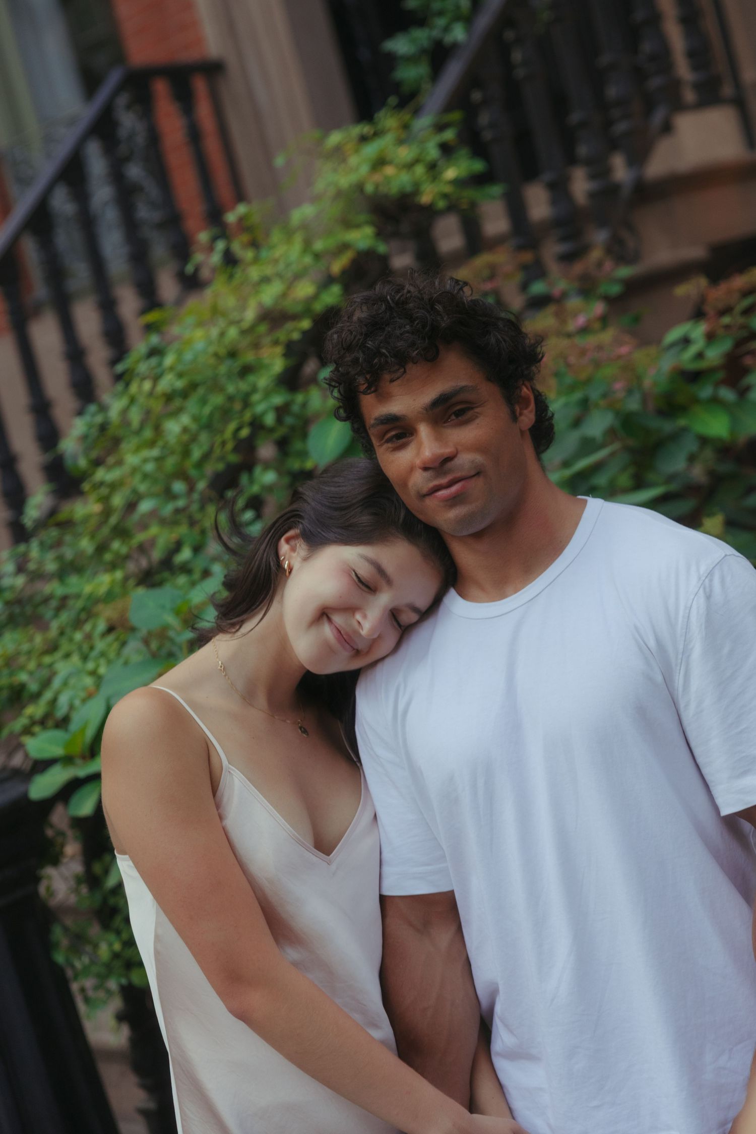 A couple embraces tenderly on an urban street with wrought iron railings and greenery in the background.
