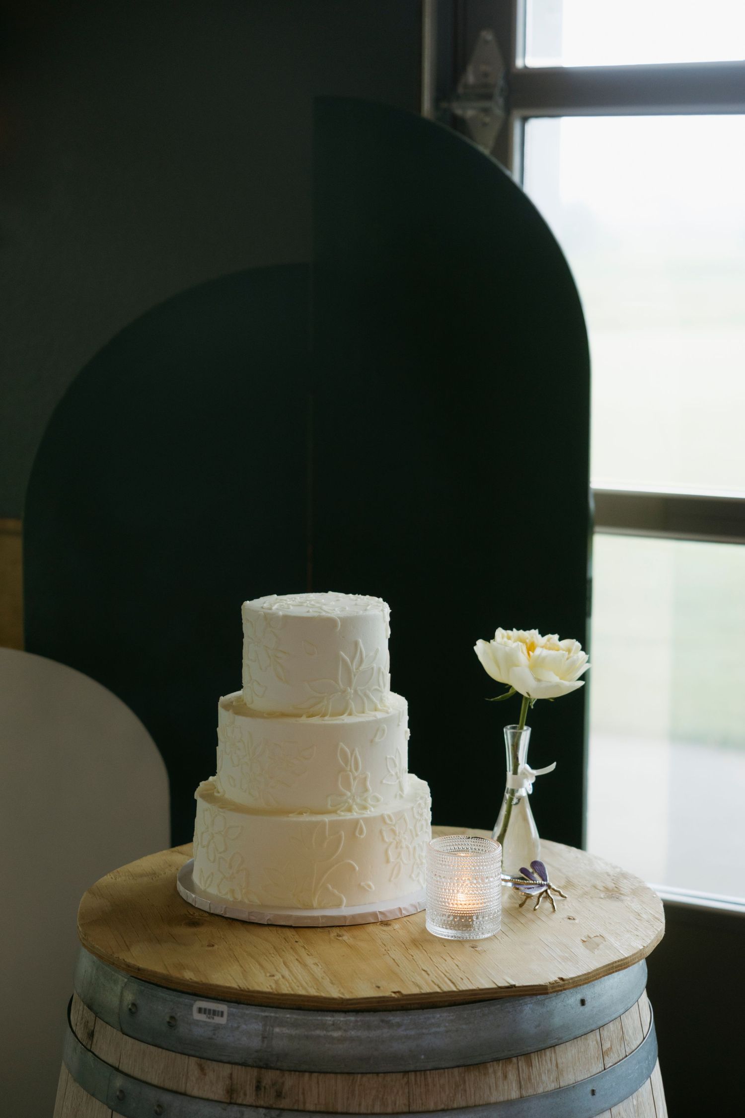 A three-tiered white wedding cake displayed on a rustic wine barrel with a single white rose in a vase beside it.