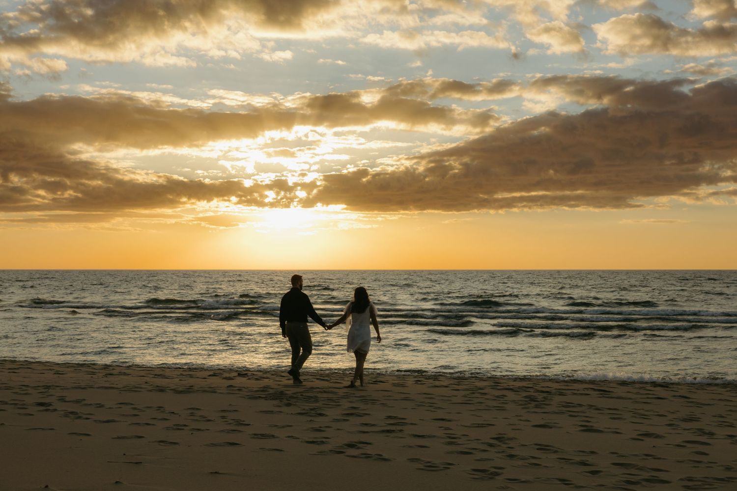 Couple holding hands walking on a sandy beach toward a stunning golden sunset over the ocean.