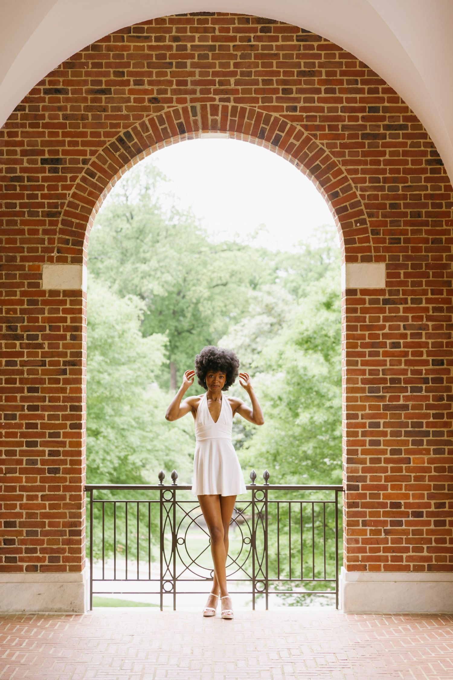 Woman in white dress stands under brick archway framed by lush greenery on sunny summer day.