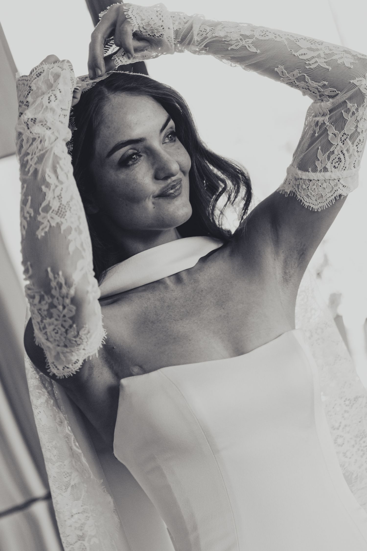 Black and white photo of a bride in lace wedding dress with arms raised, smiling elegantly.