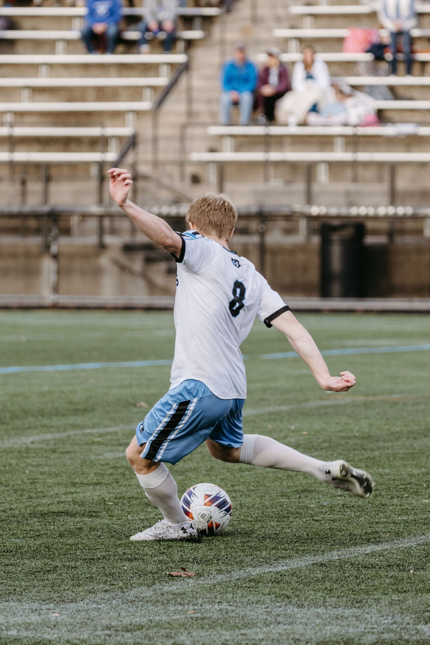 Soccer player in white jersey number 8 kicking ball on field with bleachers in background.
