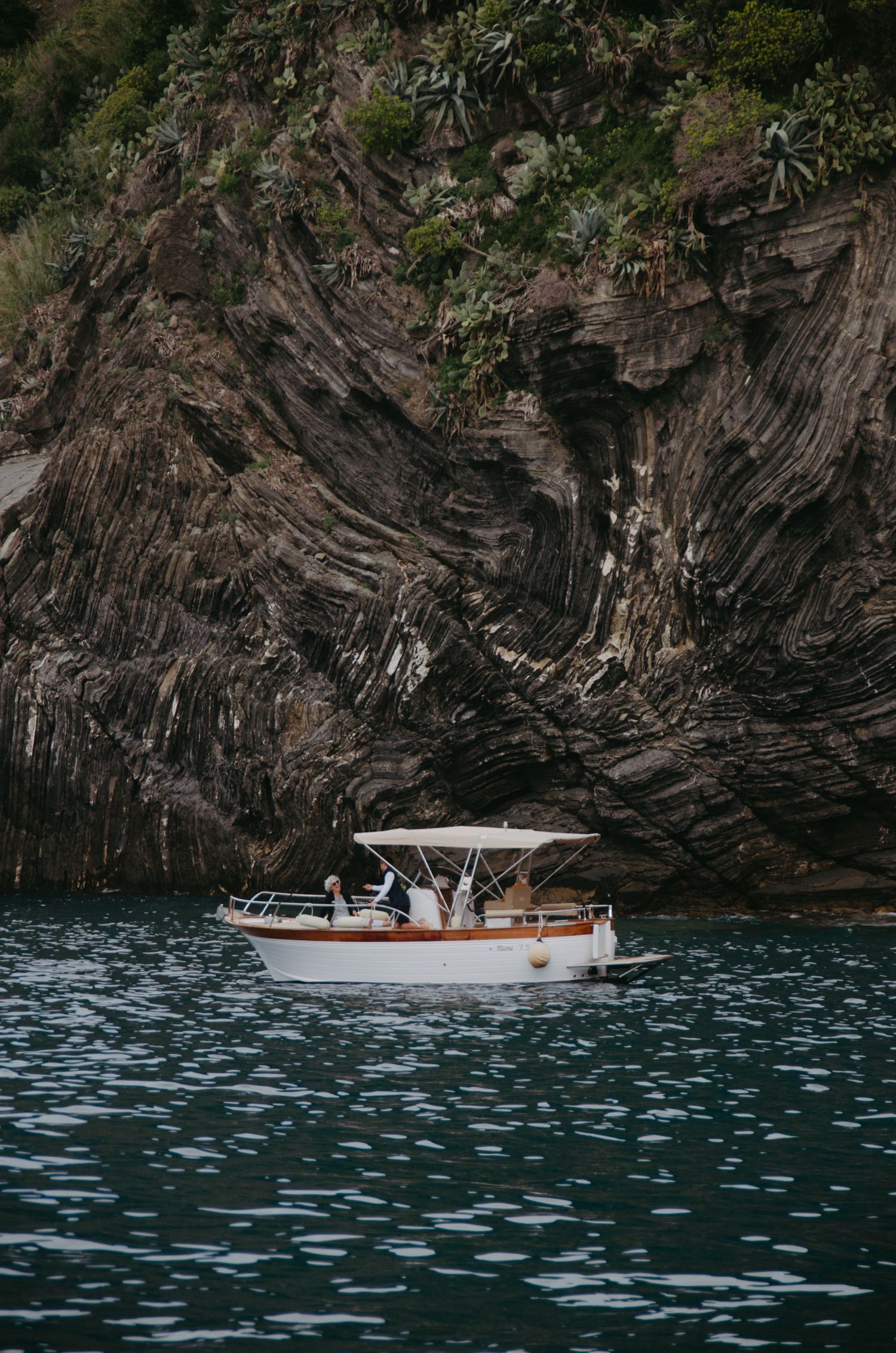 Small white boat cruises past dramatic rocky cliffs in blue Mediterranean waters.
