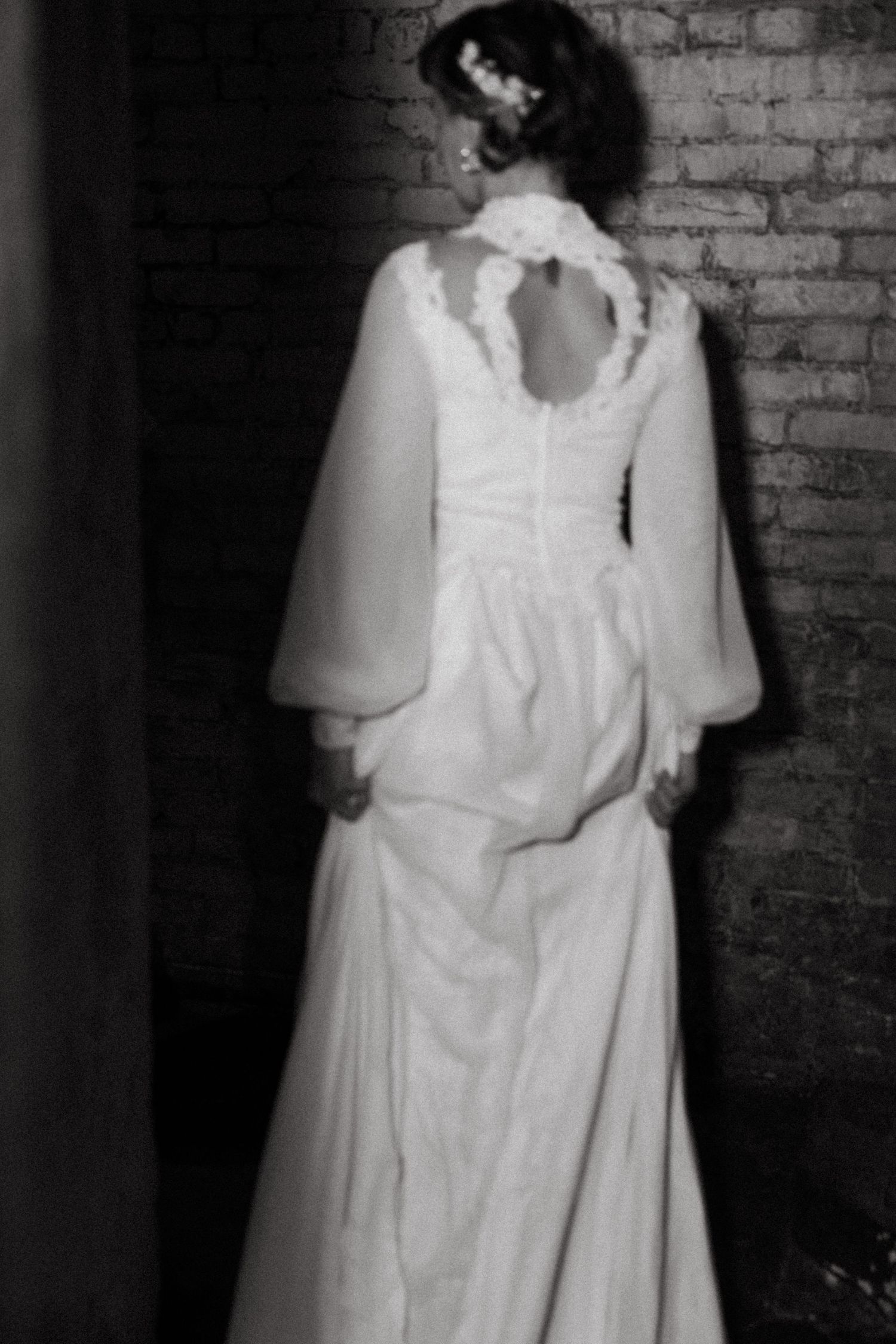 A person wearing a vintage-style white dress stands in a dark industrial hallway with exposed brick walls.