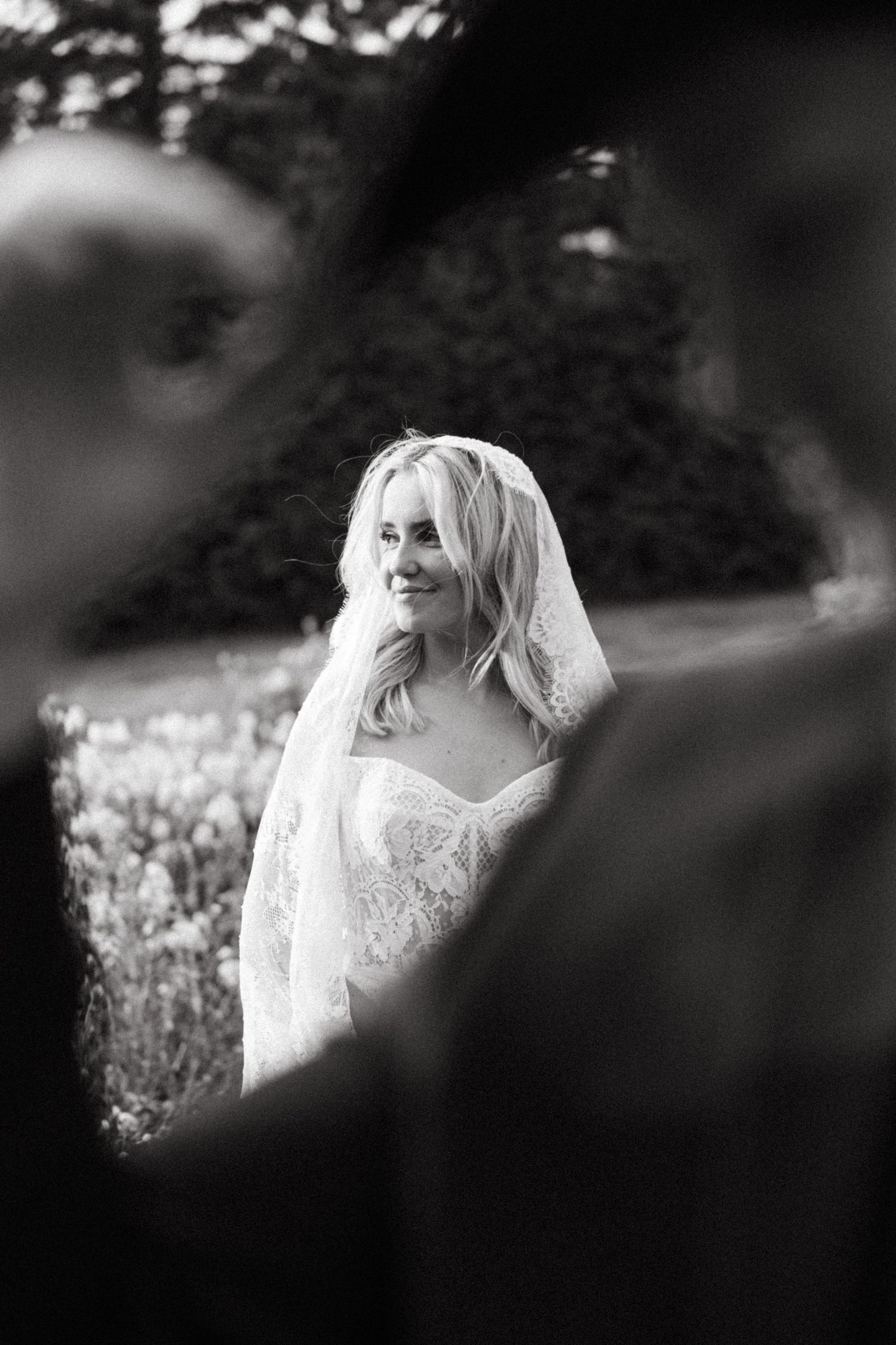 Black and white artistic portrait of a bride in a lace wedding dress and veil standing in a flower field.
